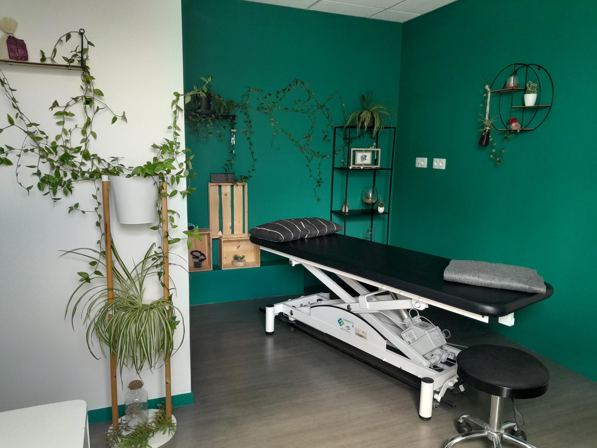 photo du cabinet de réflexologie, table devant mur en vert, des plantes un peu partout